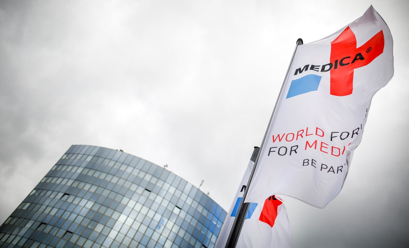 Na tle jasnego nieba widoczna jest biała powiewająca flaga z logo targów Medica 
