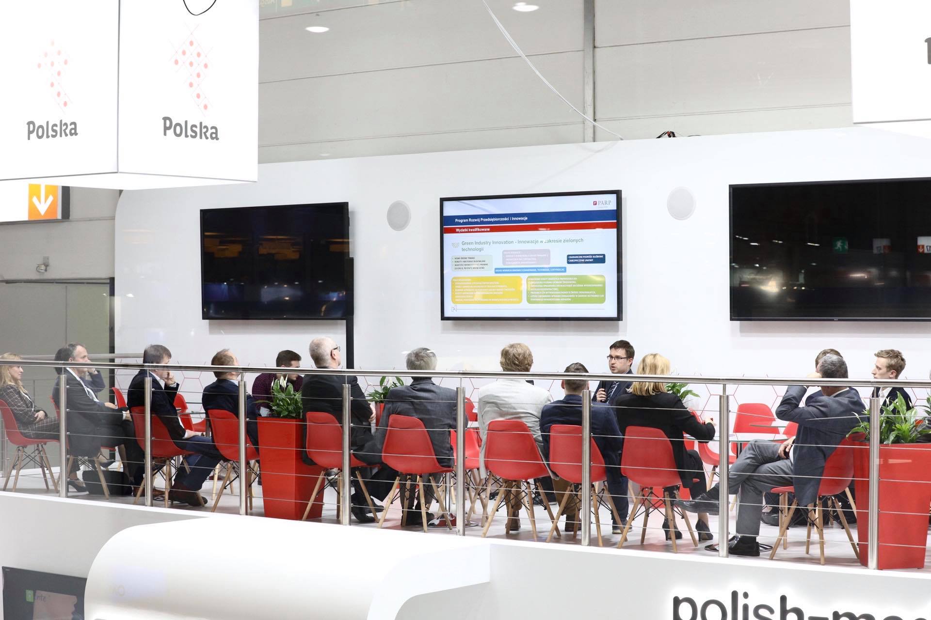 grupa ludzi w różnym wieku siedzi na czerwonych krzesełkach na antresoli. W tle widać trzy ekrany wiszące na ścianie. Z sufitu zaś zwisa sześcian z napisem Polska