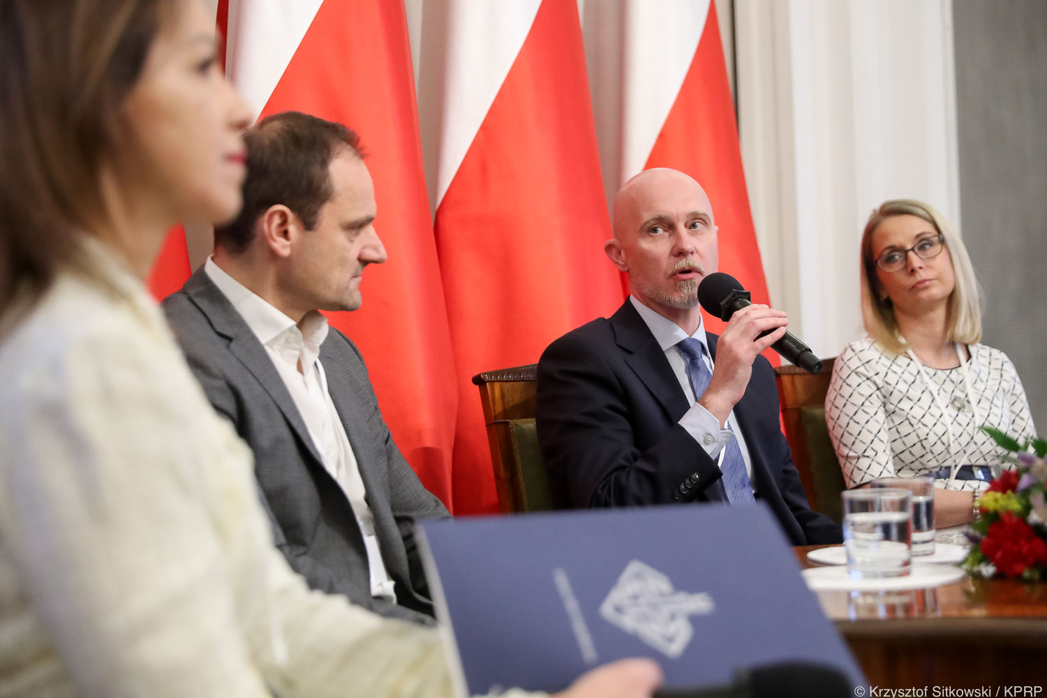Na tle polskich flag siedzą 4 osoby, jedna z nich trzyma mikrofon. 