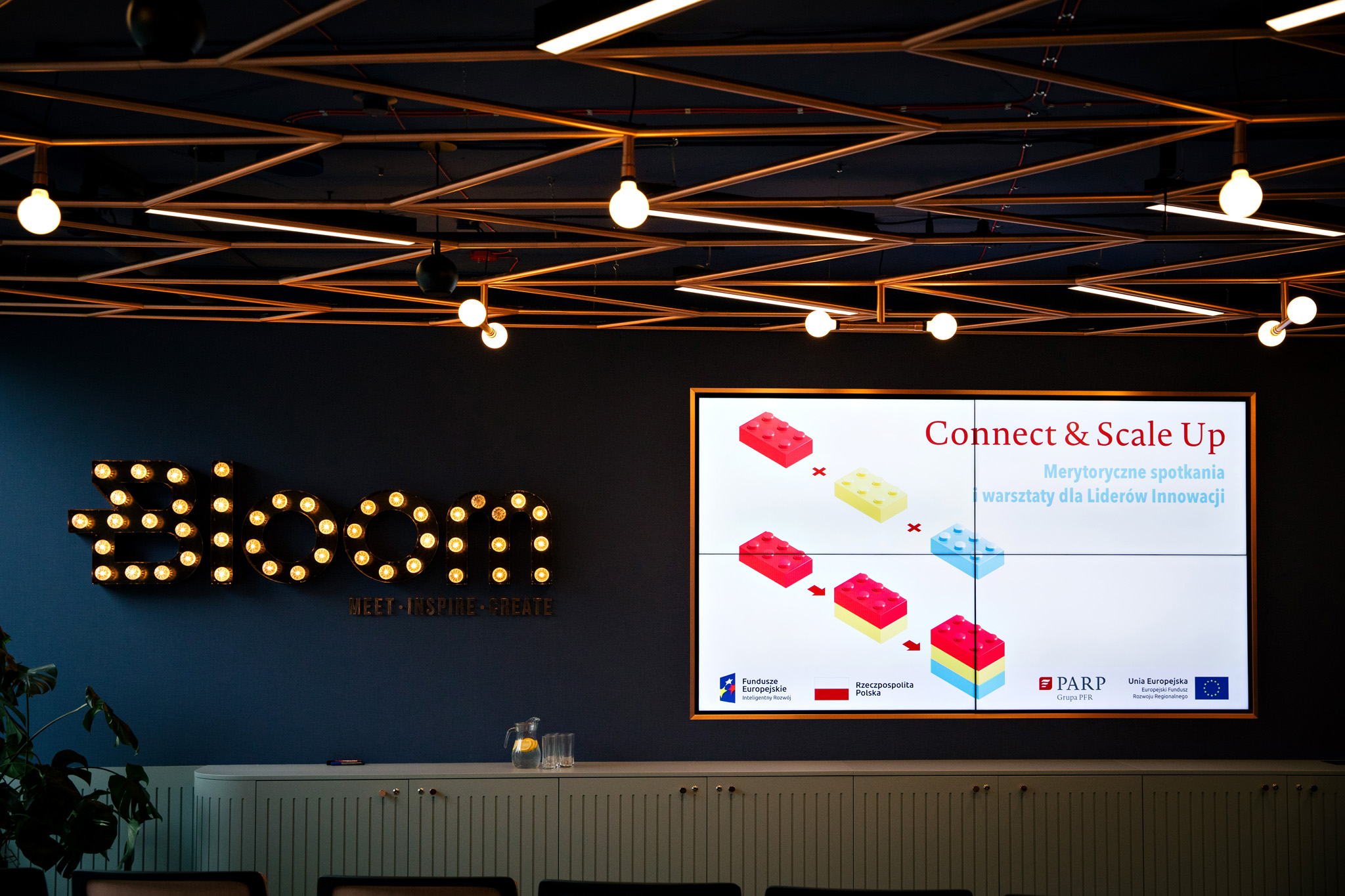 Granatowa ściana na której wyświetla się napis Bloom z żarówek. Na ekranie wiszącym na ścianie...