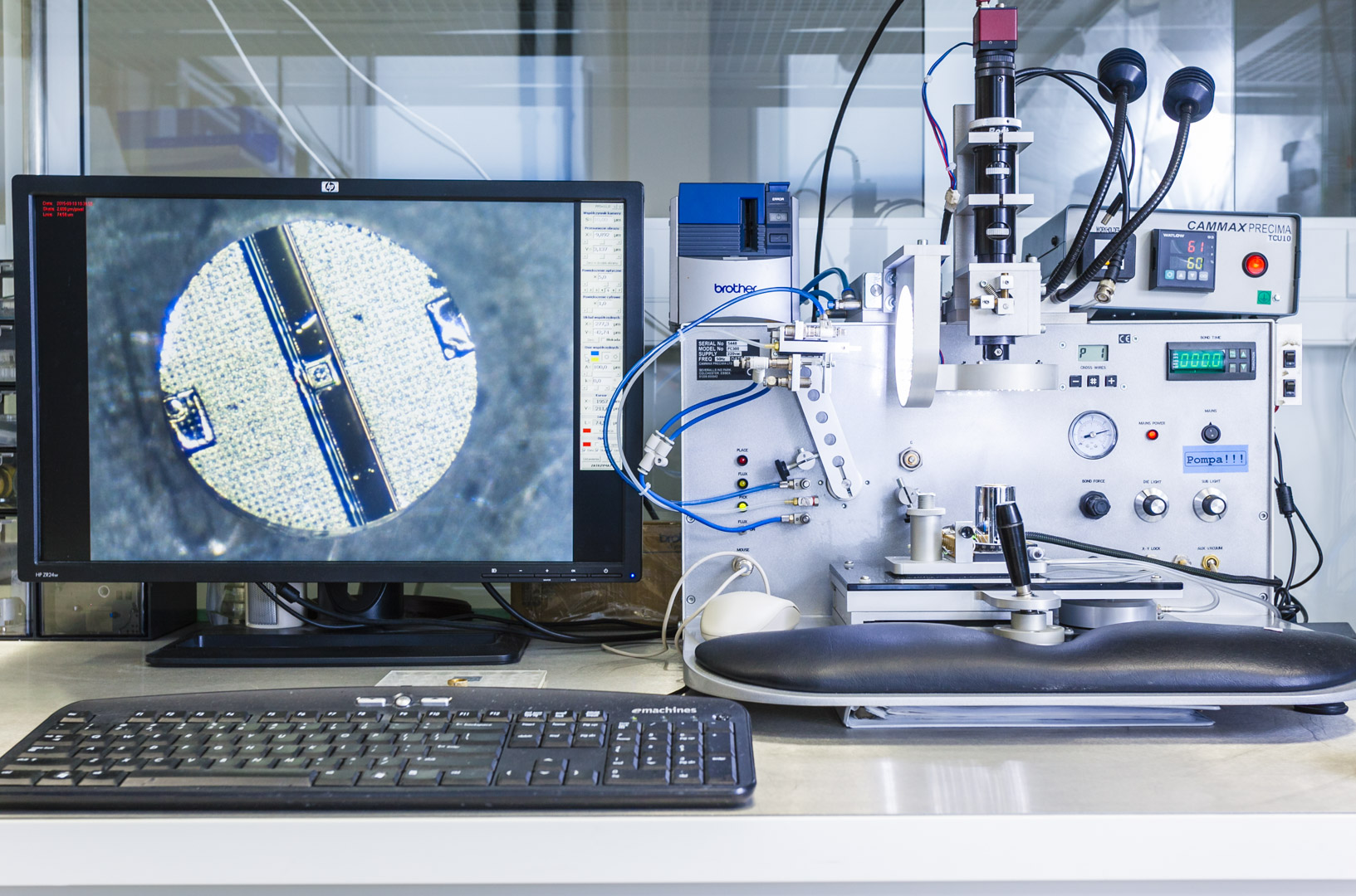 widok na monitora komputera, na którym widać zbliżenie na kolisty komponent elektroniczny. Obok monitora znajduje się specjalistyczna aparatura z diodami i pokrętłami