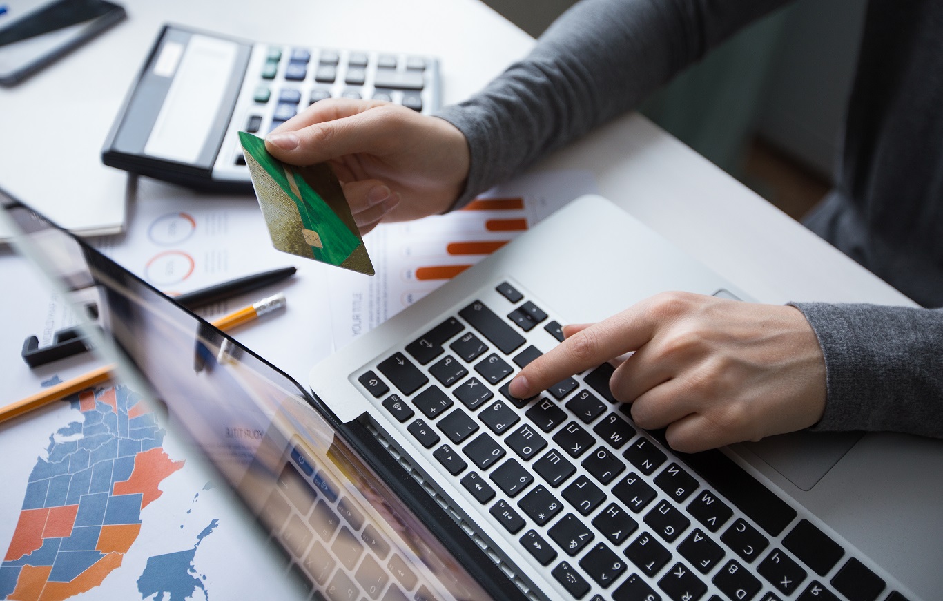 Zbliżenie na osobę siedzącą przy biurku i trzymającą w ręku kartę płatniczą. Na biurku widać też...