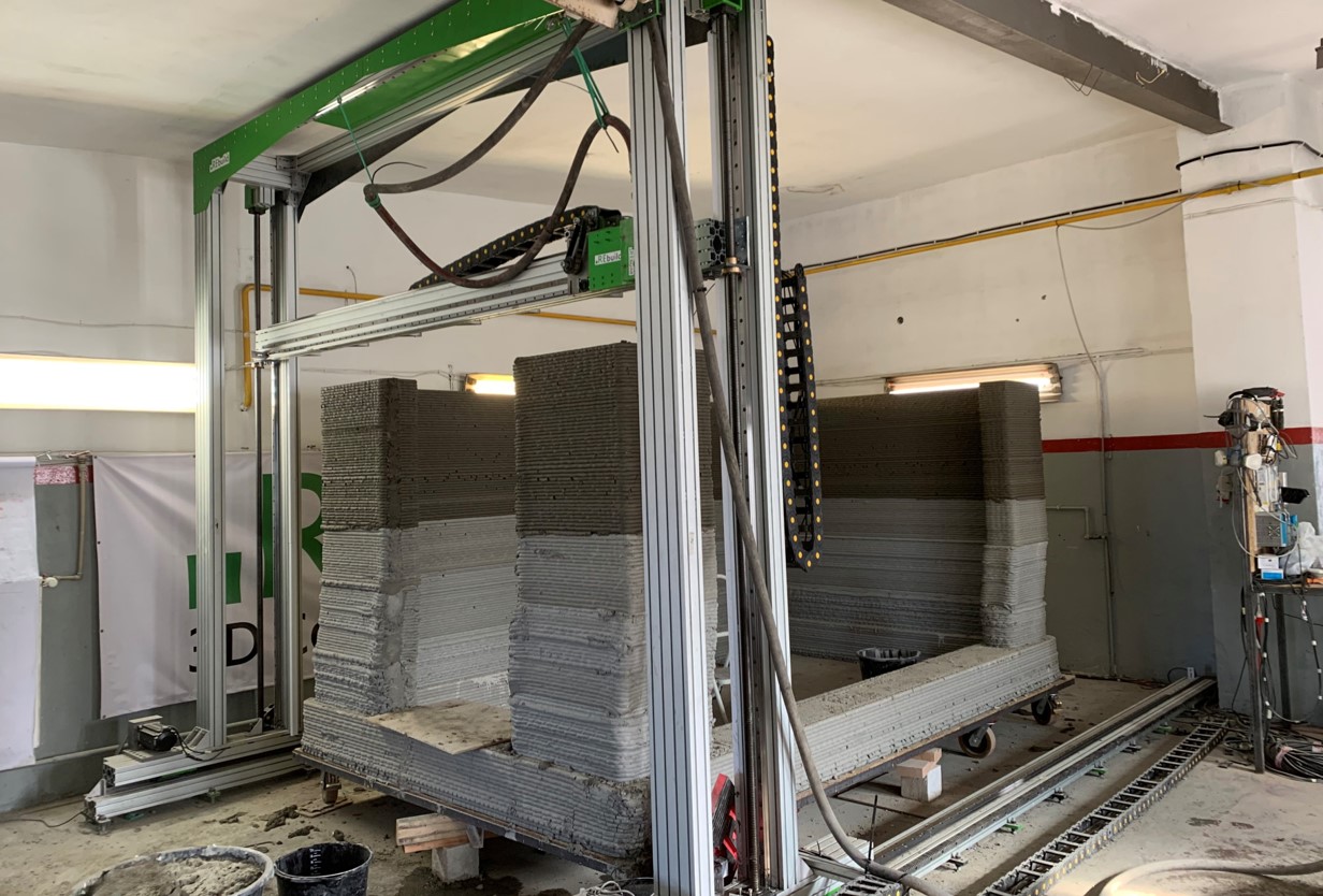 zdjęcie przedstawia hale przemysłową, w której znajduje się metalowa konstrukcja, jej rama jest przytwierdzona do sufitu oraz ściany. Obok leżących na podłodze ramion ramy widać gąsienicę. W środku ramy widać zaś drewnianą płytę, do której od spodu przytwierdzone są koła. Na drewnianej płycie znajdują się warstwy betonu tworzące ścianę.