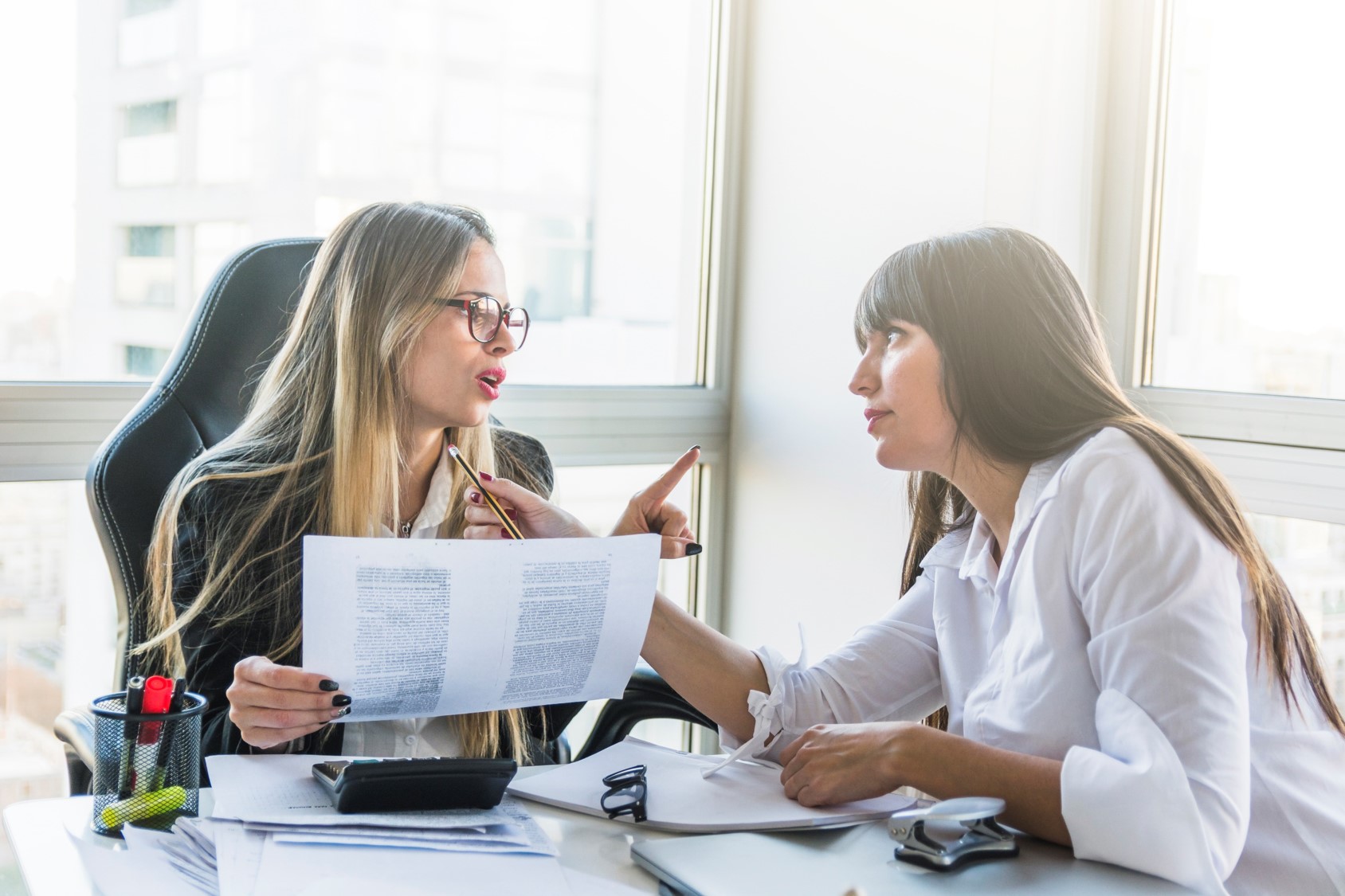 dwie młode kobiety, brunetka i blondynka, siedzą przy biurku i rozmawiają ze sobą trzymając kartki w ręku