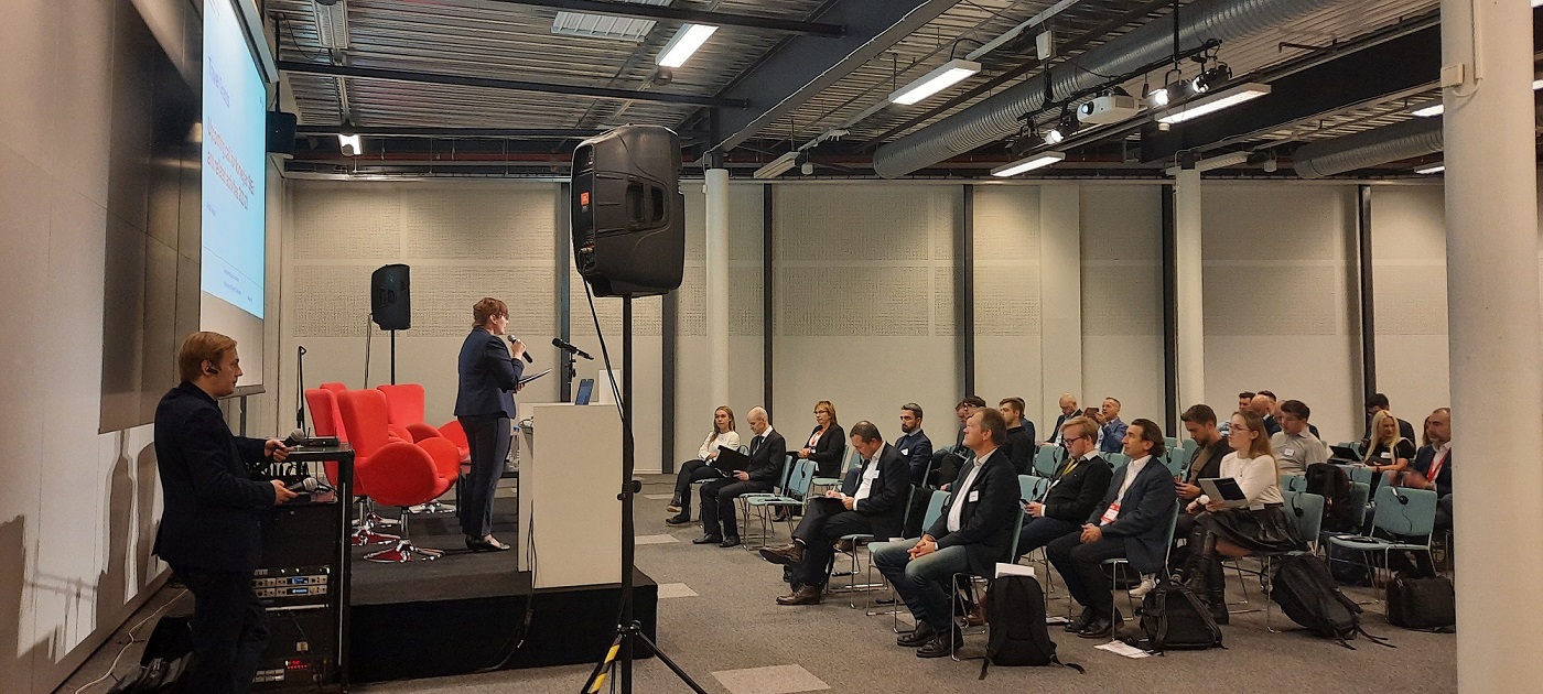 widok na salę konferencyjną z ludźmi siedzącymi na krzesłach i przysłuchującymi się wystąpieniu osoby stojącej na scenie