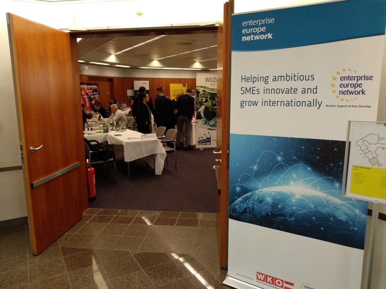 Widok na korytarz i otwarte drzwi do pomieszczenia z ludźmi, obok drzwi stoi makieta z napisem...