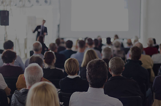 Sala konferencyjna pełna ludzi widziana od strony widowni. Prelegent prowadzi prezentację a na...