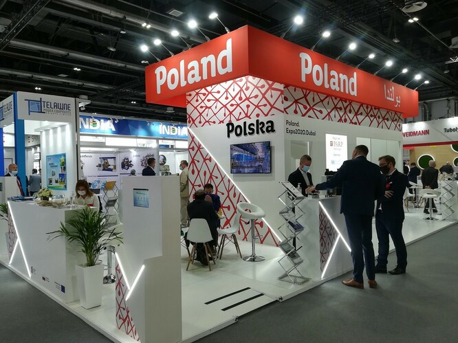 Widok na wnętrze hali wystawowej ze stoiskiem. Na górnej części kiosku widać napis "Poland". Przy...
