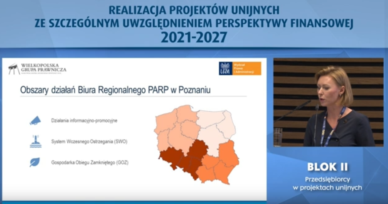 Zdjęcie przedstawia niebieski slajd podpisany –„realizacja projektów unijnych ze szczególnym uwzględnieniem perspektywy finansowej 2021-2027”. Po lewej stronie widać na białym tle napisy „obszary działania Biura Regionalnego PARP w Poznaniu: działania informacyjno-promocyjne; system wczesnego ostrzegania SWO; gospodarka obiegu zamkniętego GOZ” oraz przedstawia mapkę Polski, a po prawej stronie widać kobietę mówiącą do mikrofonu. Kobieta ubrana jest na granatowo – pod zdjęciem widać napis „Blok II Przedsiębiorcy w projektach unijnych”.