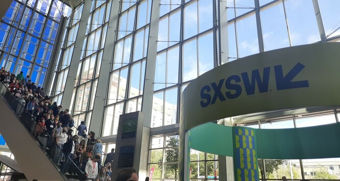 Widok na przeszklony hol i schody ruchome, na których widać ludzi. Nad wejściem do budynku widać...